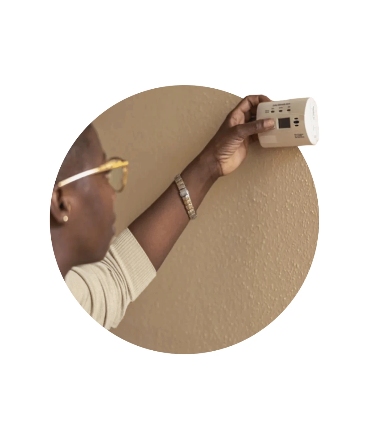 A person wearing glasses holds an electronic device against a beige wall, possibly using a stud finder or similar tool.